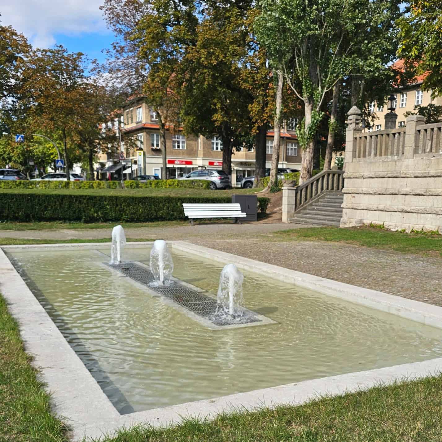 Ein Brunnen mit drei kleinen Fontänen in einer Grünanlage. Im Hintergrund eine weiße Parkbank und eine Steintreppe.
