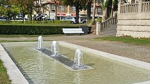 Ein Brunnen mit drei kleinen Fontänen in einer Grünanlage. Im Hintergrund eine weiße Parkbank und eine Steintreppe.