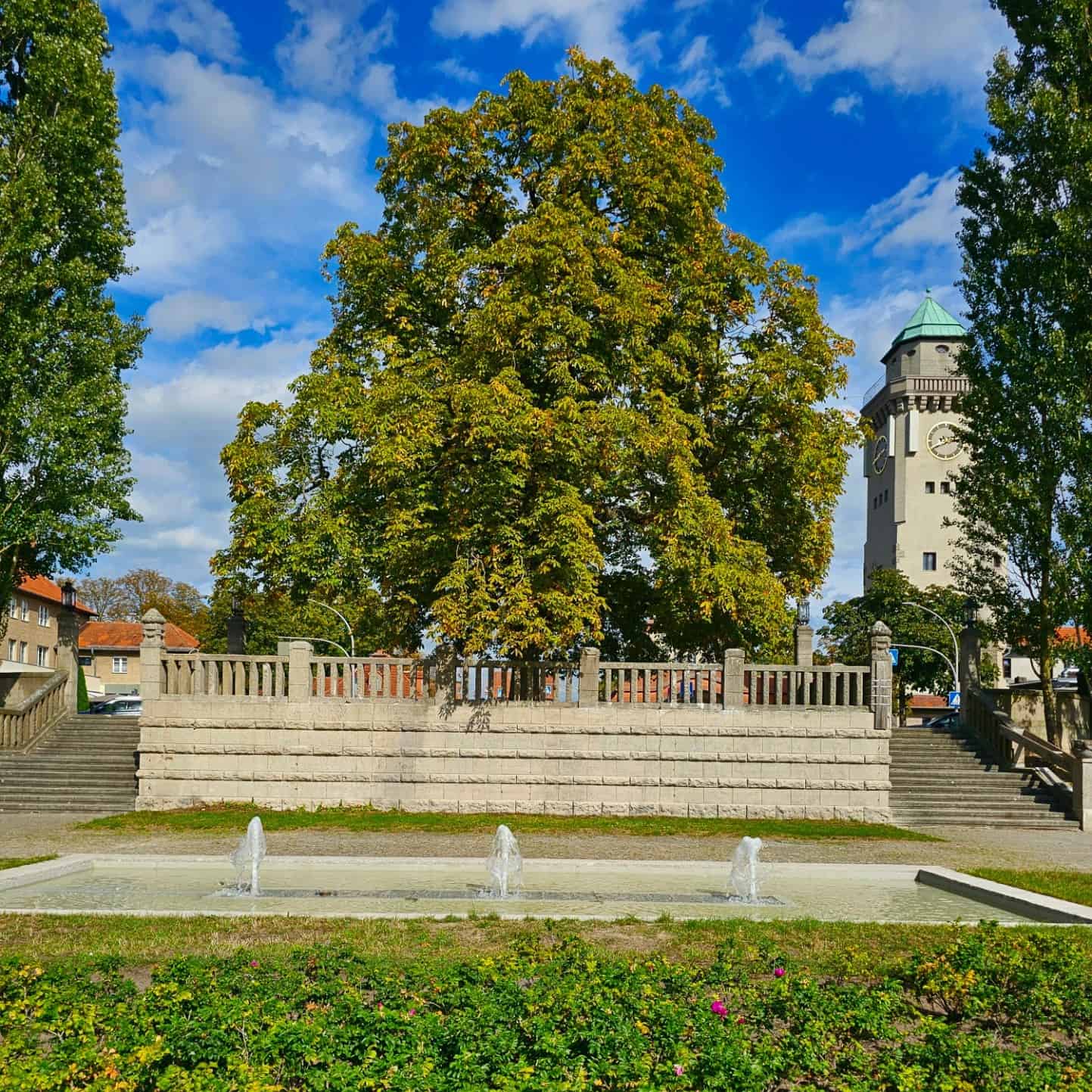 Ein Brunnen vor einer Treppenanlage mit steinerner Brüstung und einem großen kastanienbaum. Im Hintergrund der Frohnauer Kasinoturm.