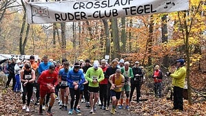 Läuferinnen und Läufer laufen unter einem Banner mit der Aufschrift Crosslauf Ruder-Club Tegel los, ringsum herbstlich gefärbter Wald und Laub auf dem Weg.