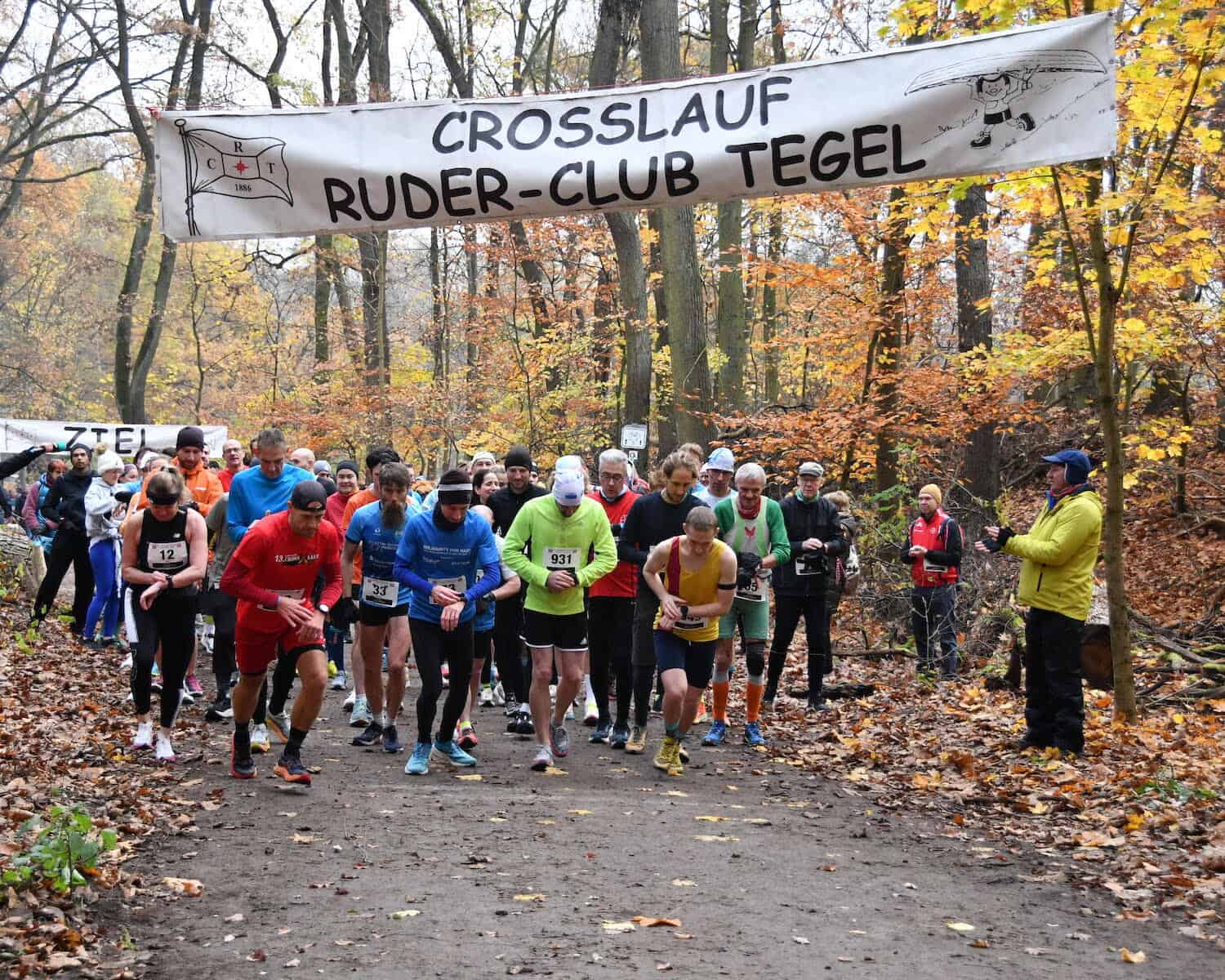 Läuferinnen und Läufer laufen unter einem Banner mit der Aufschrift Crosslauf Ruder-Club Tegel los, ringsum herbstlich gefärbter Wald und Laub auf dem Weg.