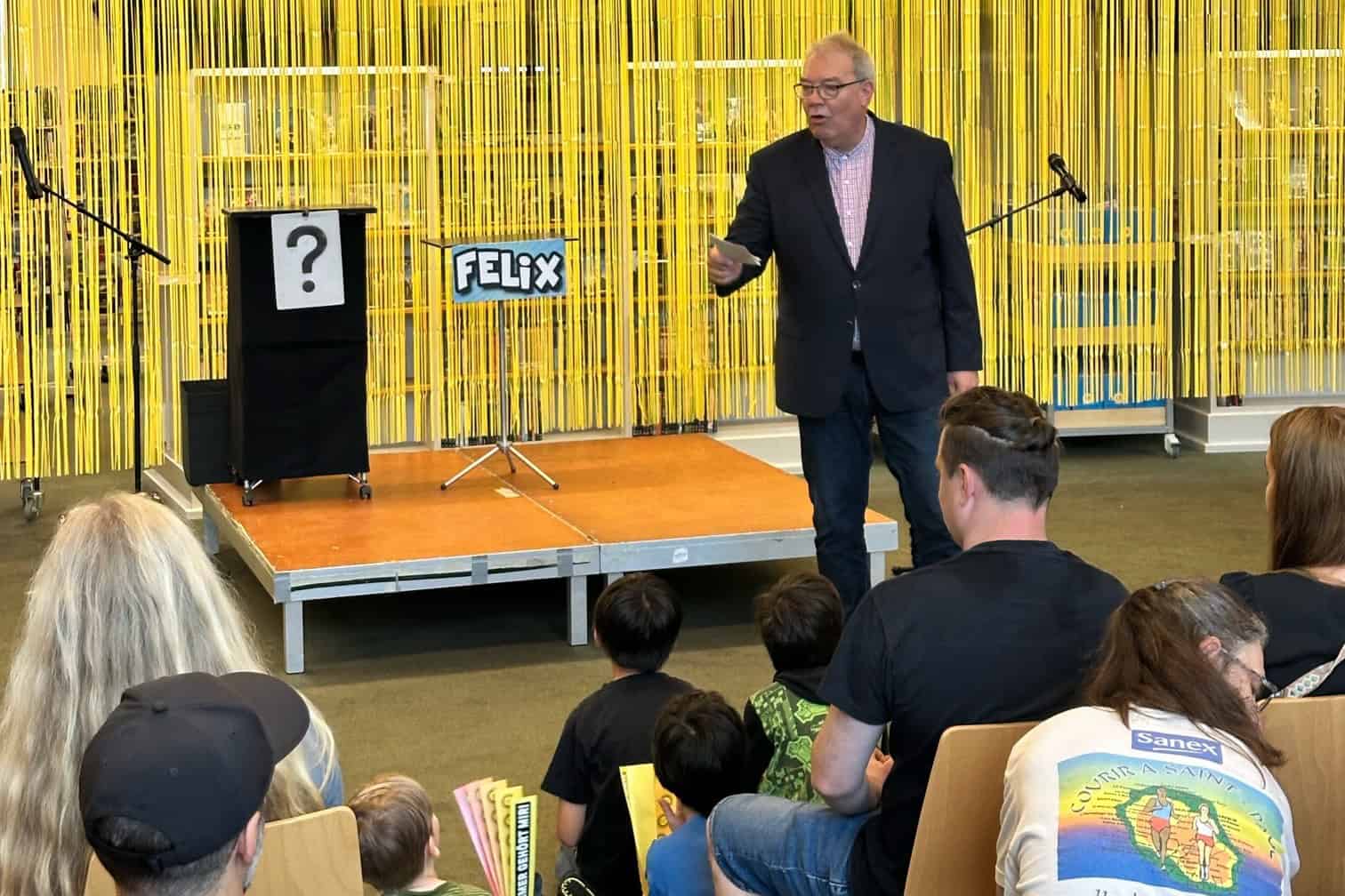 Kulturstadtrat Harald Muschner steht vor einer gelben Dekowand und spricht zu sitzenden Kindern und Erwachsenen in der Humboldt-Bibliothek.
