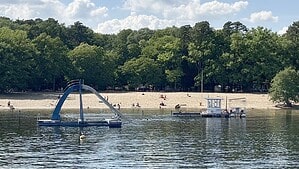 Blick über das Wasser des Tegeler Sees auf den Sandstrand des Strandbads Tegel mit Wasserrutsche und Badeinseln vor grünen Bäumen.
