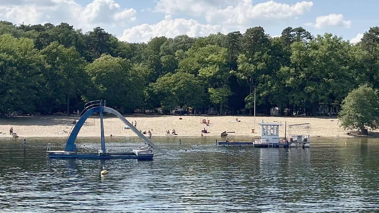 Blick über das Wasser des Tegeler Sees auf den Sandstrand des Strandbads Tegel mit Wasserrutsche und Badeinseln vor grünen Bäumen.
