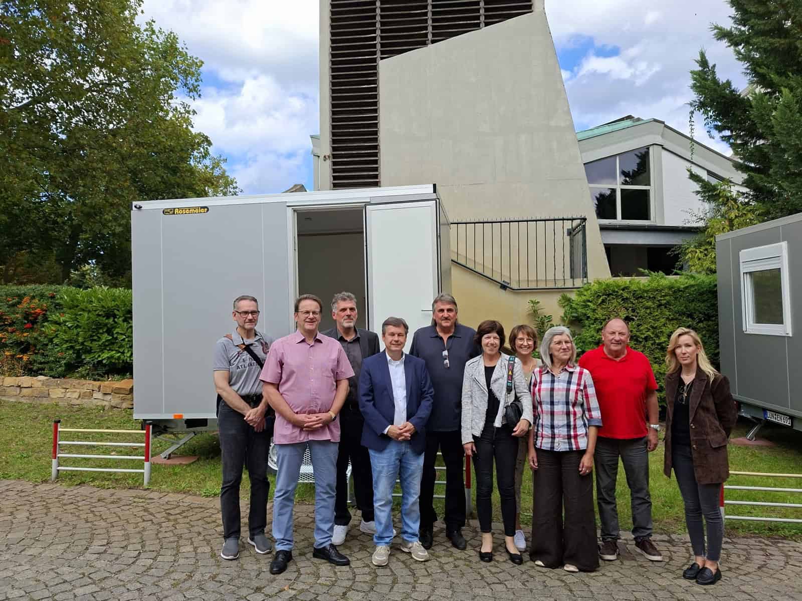 Elf Personen stehen vor einem weißen Tiny House auf einem Anhänger, dessen Tür offen steht; rechts im Bild ein zweiter Wagen.