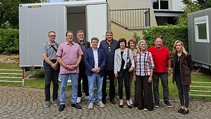 Elf Personen stehen vor einem weißen Tiny House auf einem Anhänger, dessen Tür offen steht; rechts im Bild ein zweiter Wagen.