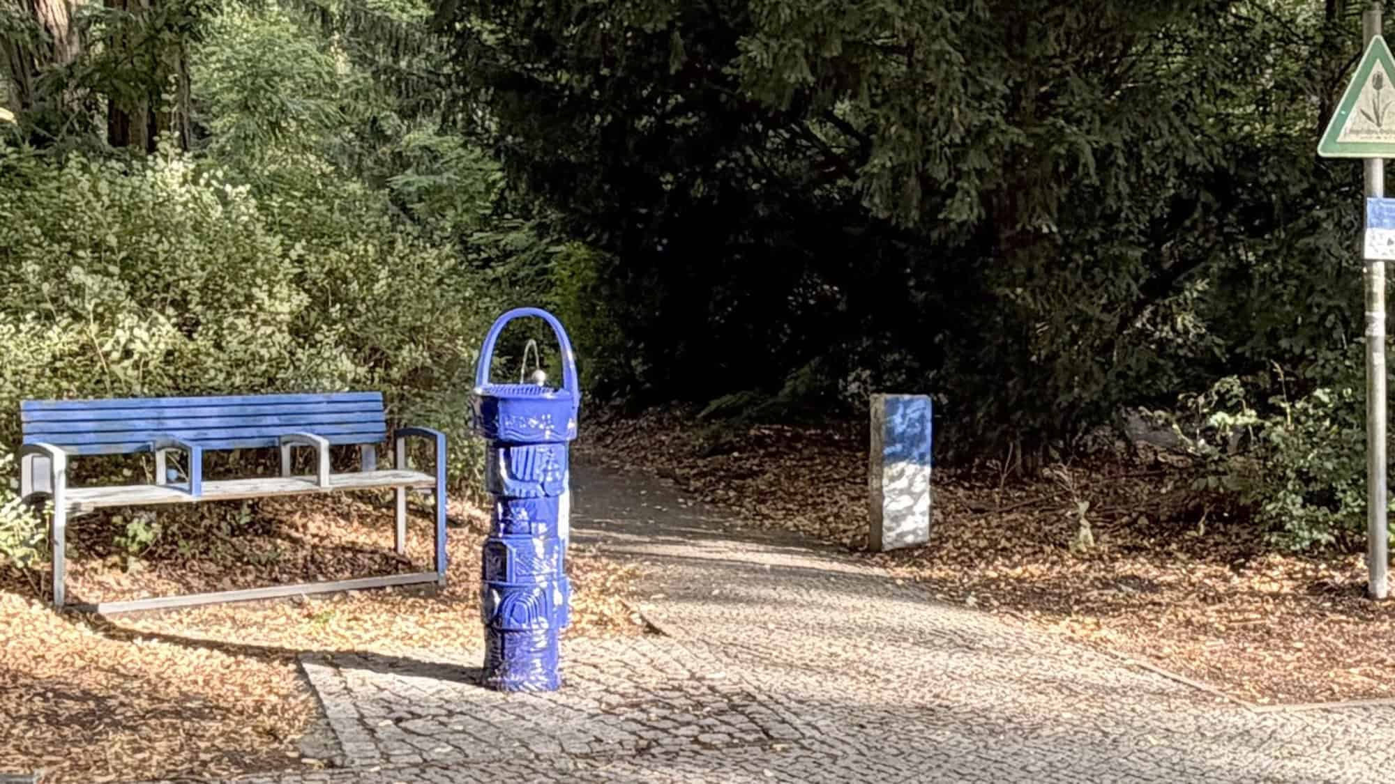 Ein Trinkbrunnen steht vor einer Parkbank am Rande einer Parkanlage.