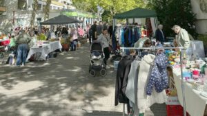 Trödelmarkt unter Bäumen auf einem gepflasterten Weg mit Ständen, Kleiderständern und Pavillons, dazwischen Besucher mit Kinderwagen, im Hintergrund ein mehrstöckiges Wohngebäude