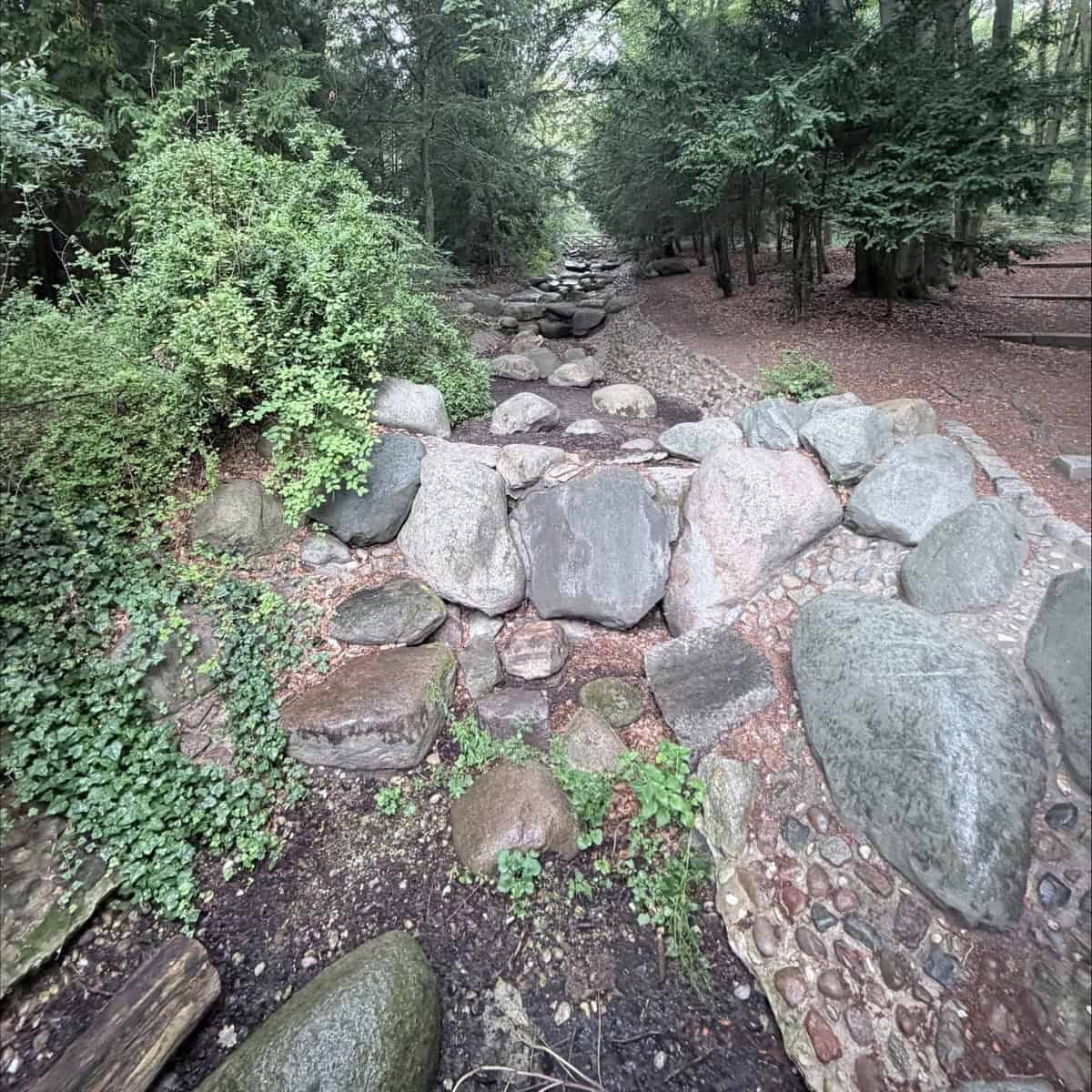 Trockenes Bachbett aus Findlingen und Feldsteinen im Steinbergpark, dahinter hohe Bäume