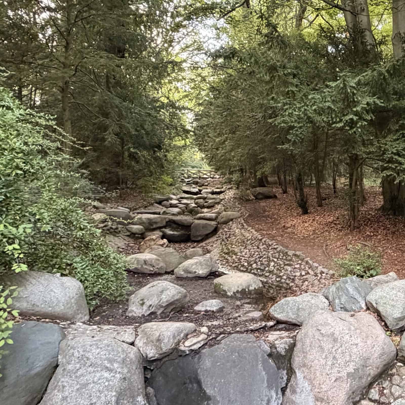 Trockenes, steinernes Bachbett eines künstlichen Wasserfalls, zwischen den Findlingen wachsen Gräser.