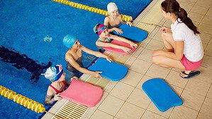 Symbolbild: Vier Kinder mit Badekappen und Schwimmbrettern halten sich am Beckenrand fest und hören einer Trainerin zu, die am Rand hockt.