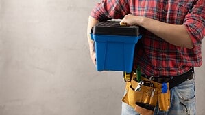 Mann im Karohemd mit Werkzeuggürtel hält eine blaue Werkzeugkiste vor einer grauen Wand