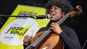 Eine Sängerin mit Lockenkopf singt in ein Mikrofon und spielt dabei Cello, hinter ihr ein gelbes Banner mit der Aufschrift Jazz & Soul.