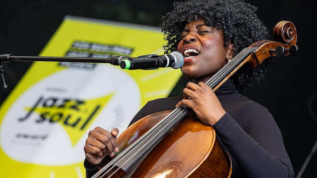 Eine Sängerin mit Lockenkopf singt in ein Mikrofon und spielt dabei Cello, hinter ihr ein gelbes Banner mit der Aufschrift Jazz & Soul.