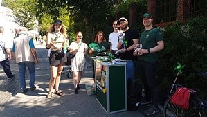 Sechs Personen stehen an einem grünen Infostand auf einem Gehweg, mehrere halten Flyer und grüne Fähnchen in der Hand.