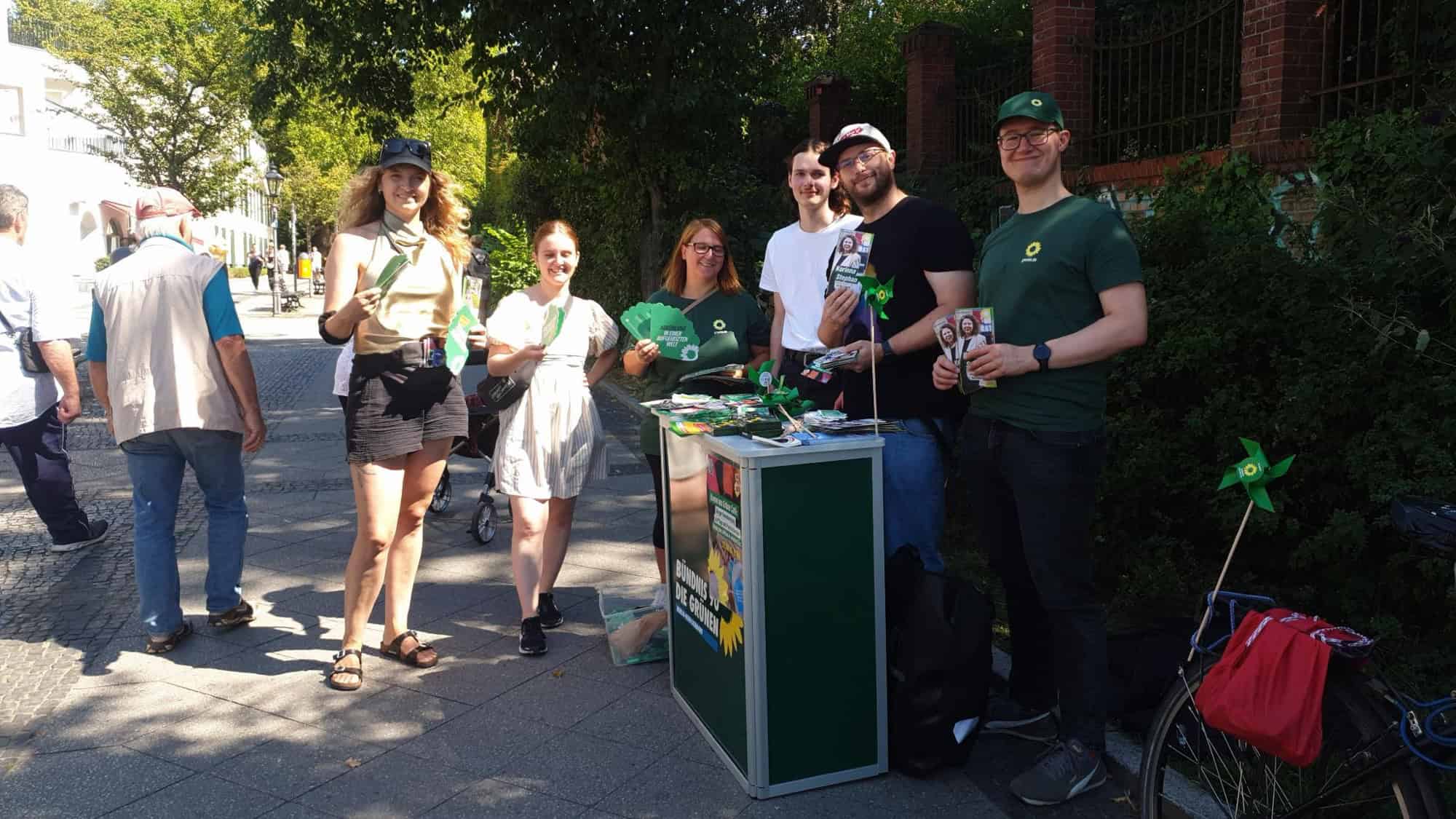 Sechs Personen stehen an einem grünen Infostand auf einem Gehweg, mehrere halten Flyer und grüne Fähnchen in der Hand.