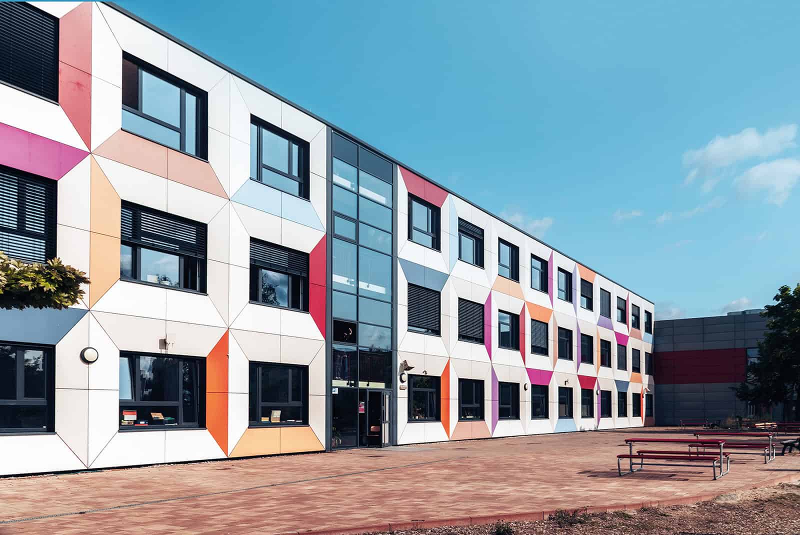 Weißes, dreigeschossiges Schulgebäude mit bunten Farbflächen an der Fassade und Bänken auf dem gepflasterten Vorplatz.