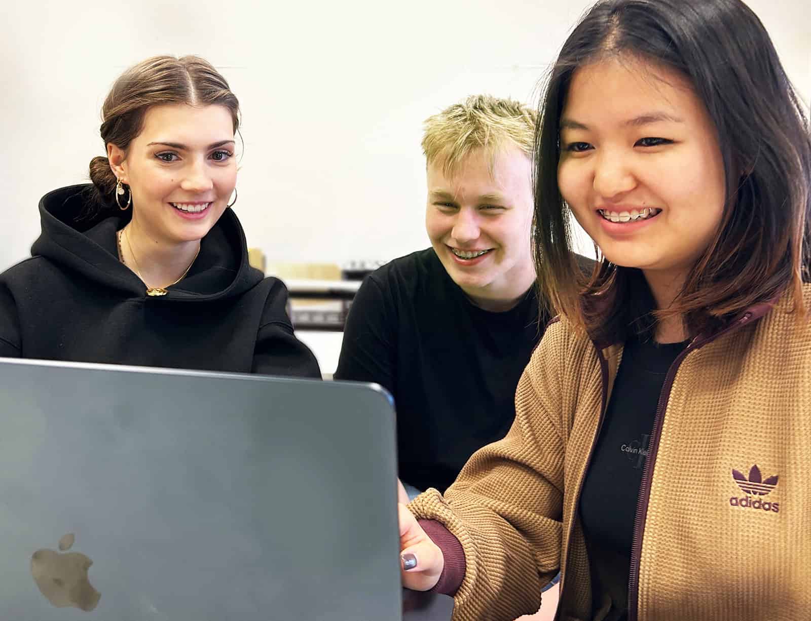 Zwei Schülerinnen und ein Schüler sitzen nebeneinander vor einem aufgeklappten Laptop und lächeln.