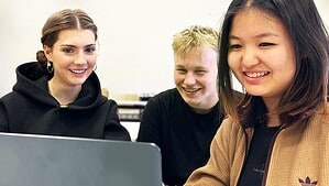 Zwei Schülerinnen und ein Schüler sitzen nebeneinander vor einem aufgeklappten Laptop und lächeln.