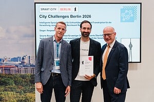 Drei Männer stehen vor einem Bildschirm mit der Aufschrift City Challenges Berlin 2026, der mittlere hält eine Urkunde.
