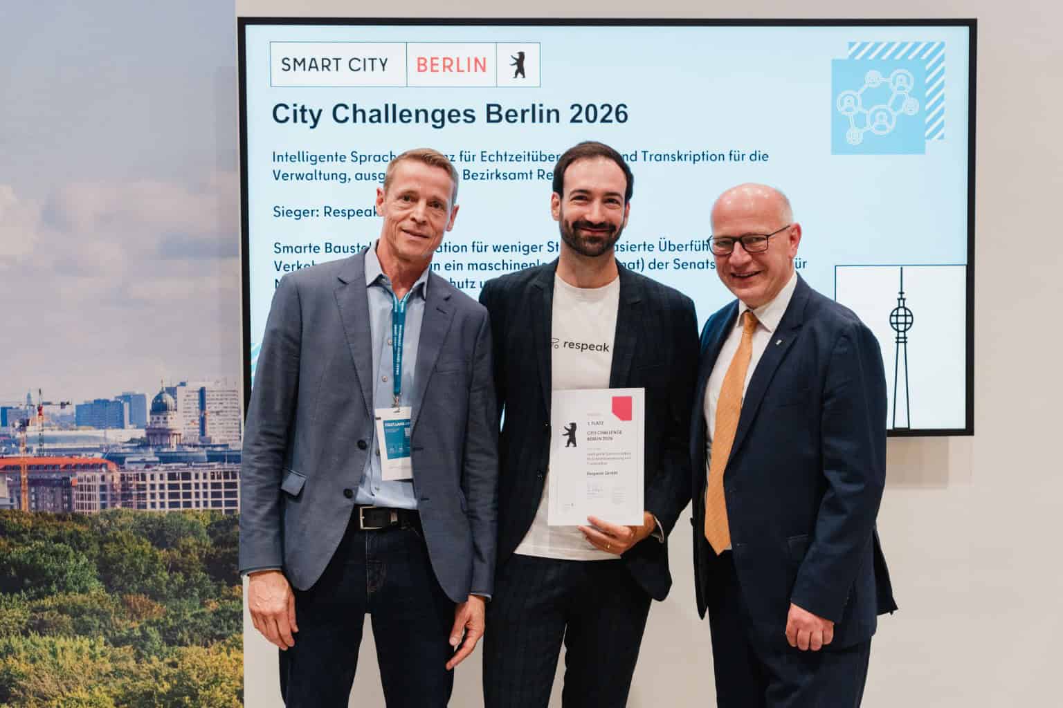 Drei Männer stehen vor einem Bildschirm mit der Aufschrift City Challenges Berlin 2026, der mittlere hält eine Urkunde.