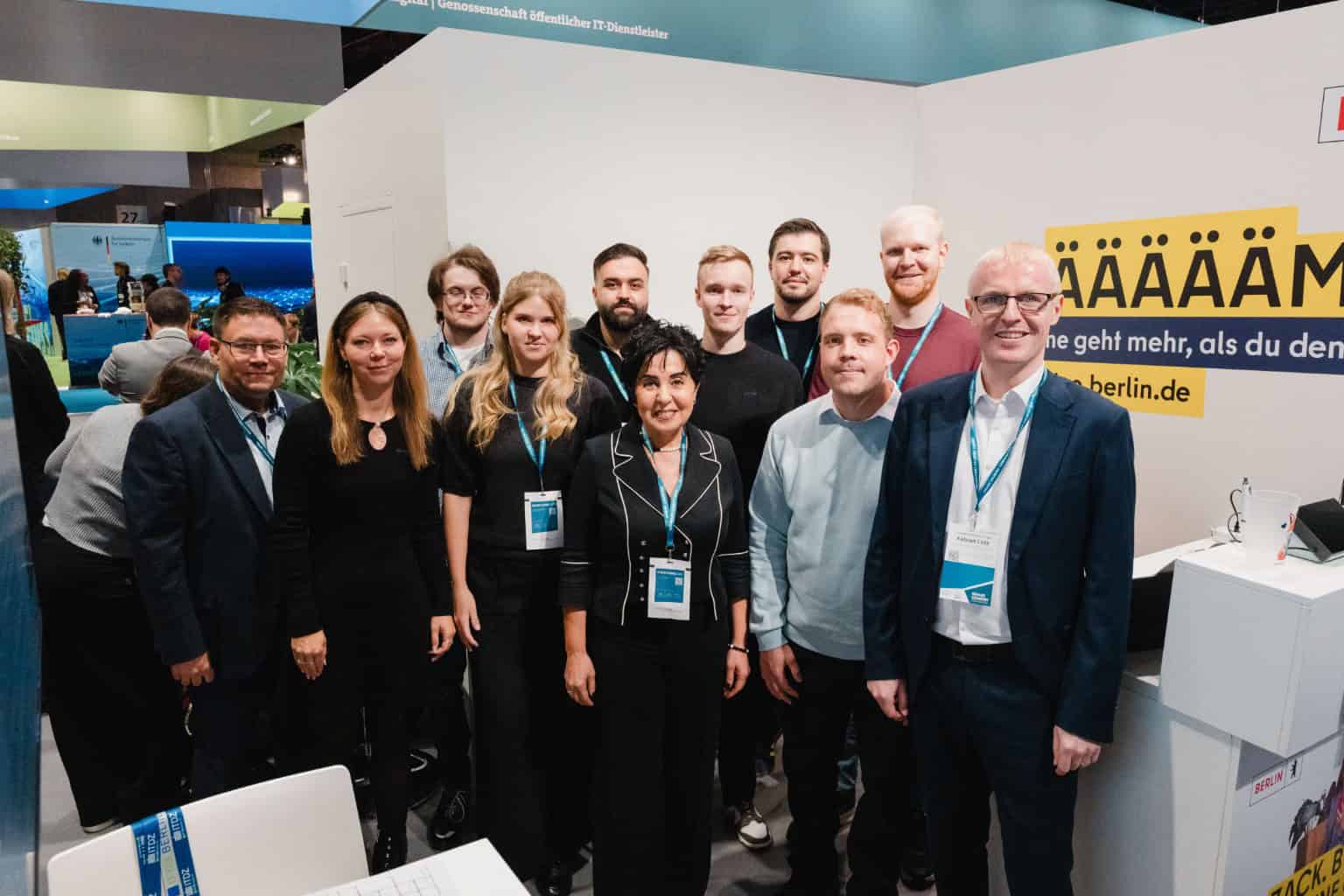 Gruppenfoto von zwölf Personen mit Konferenzausweisen an einem Messestand, in der Mitte vorne eine Frau mit dunklen Haaren.