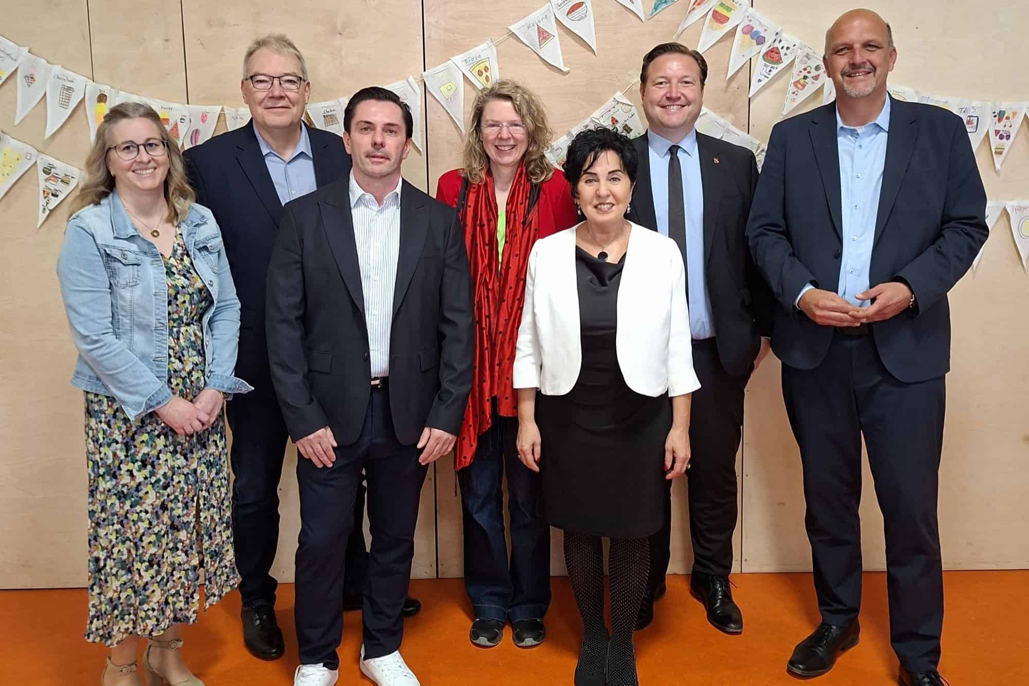 Sieben Personen aus Bezirksamt, Senat und Schulleitung stehen vor einer Holzwand mit bemalten Wimpelketten