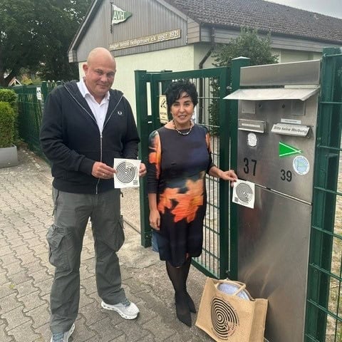 Ein Mann und eine Frau bringen am Tor des Angelvereins Heiligensee einen runden Sticker an