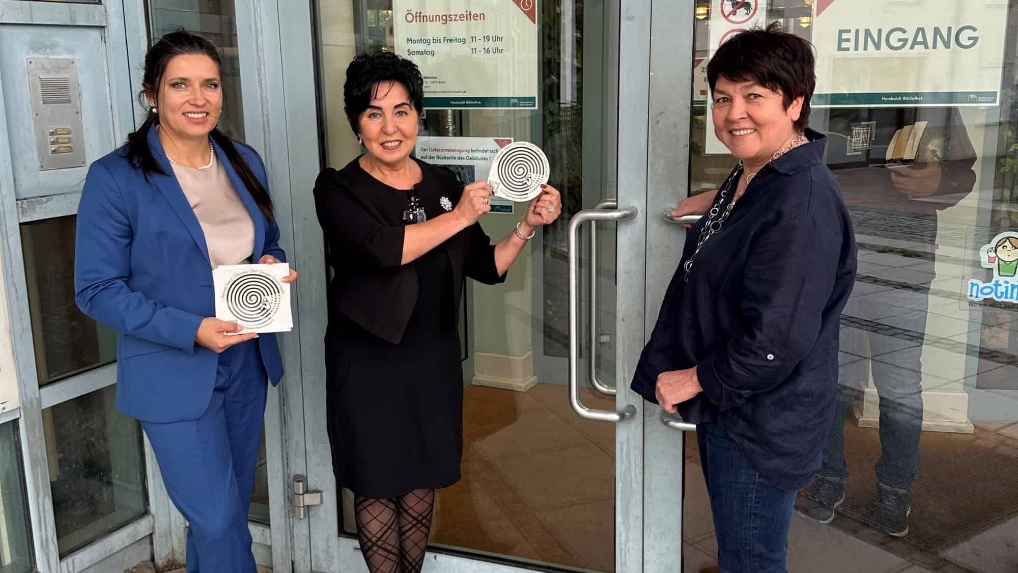 Drei Frauen halten am Glaseingang der Humboldt-Bibliothek runde Sticker mit Spiralmotiv