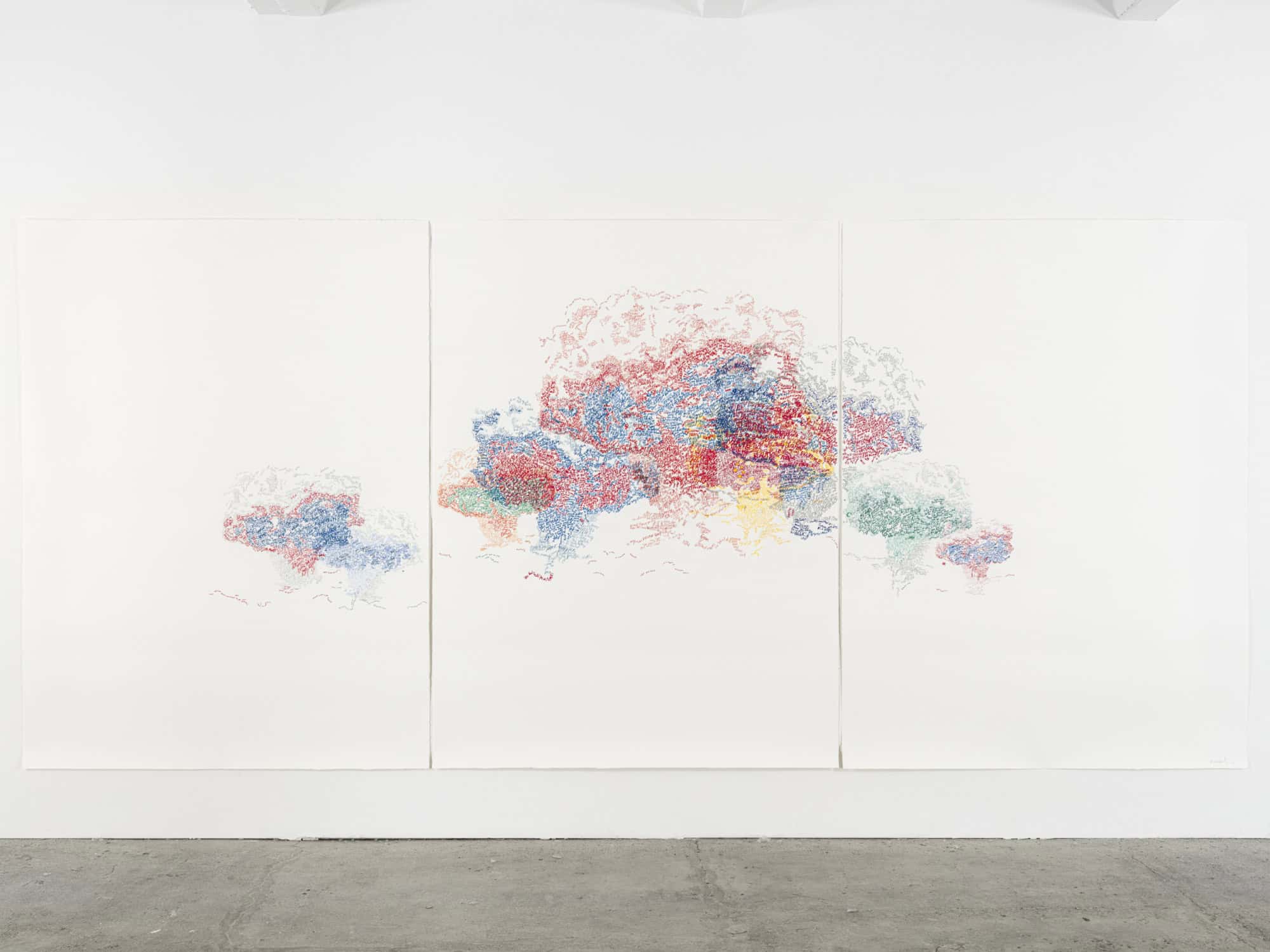 Großes dreiteiliges Werk auf weißem Papier an einer Galeriewand, in der Mitte eine dichte Wolke aus roten, blauen, grünen und gelben Schriftzeichen.