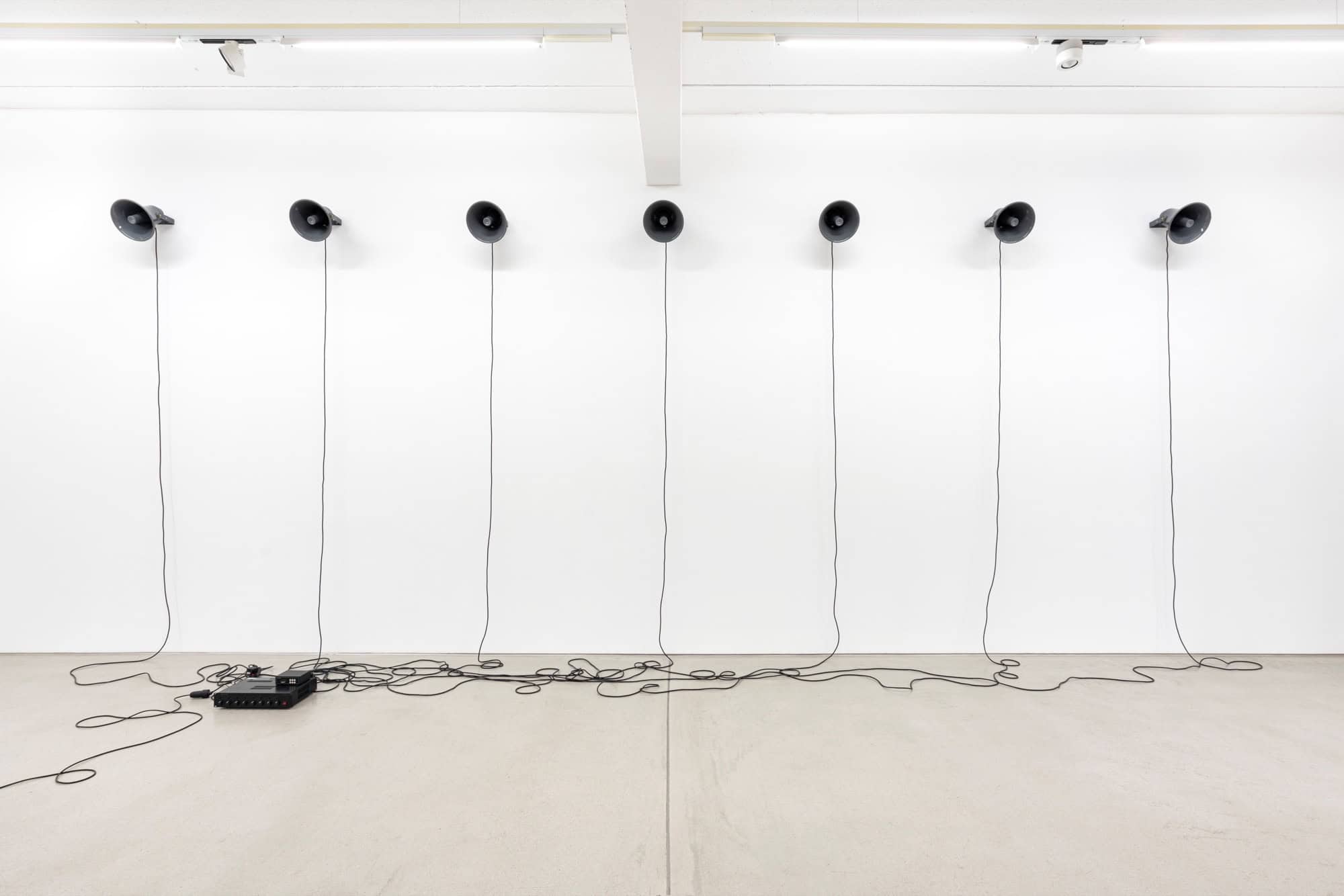 Sieben schwarze Trichterlautsprecher hängen in einer Reihe an einer weißen Wand, ihre Kabel laufen auf dem hellen Boden zu einem Verstärker zusammen.