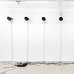 Sieben schwarze Trichterlautsprecher hängen in einer Reihe an einer weißen Wand, ihre Kabel laufen auf dem hellen Boden zu einem Verstärker zusammen.