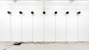 Sieben schwarze Trichterlautsprecher hängen in einer Reihe an einer weißen Wand, ihre Kabel laufen auf dem hellen Boden zu einem Verstärker zusammen.