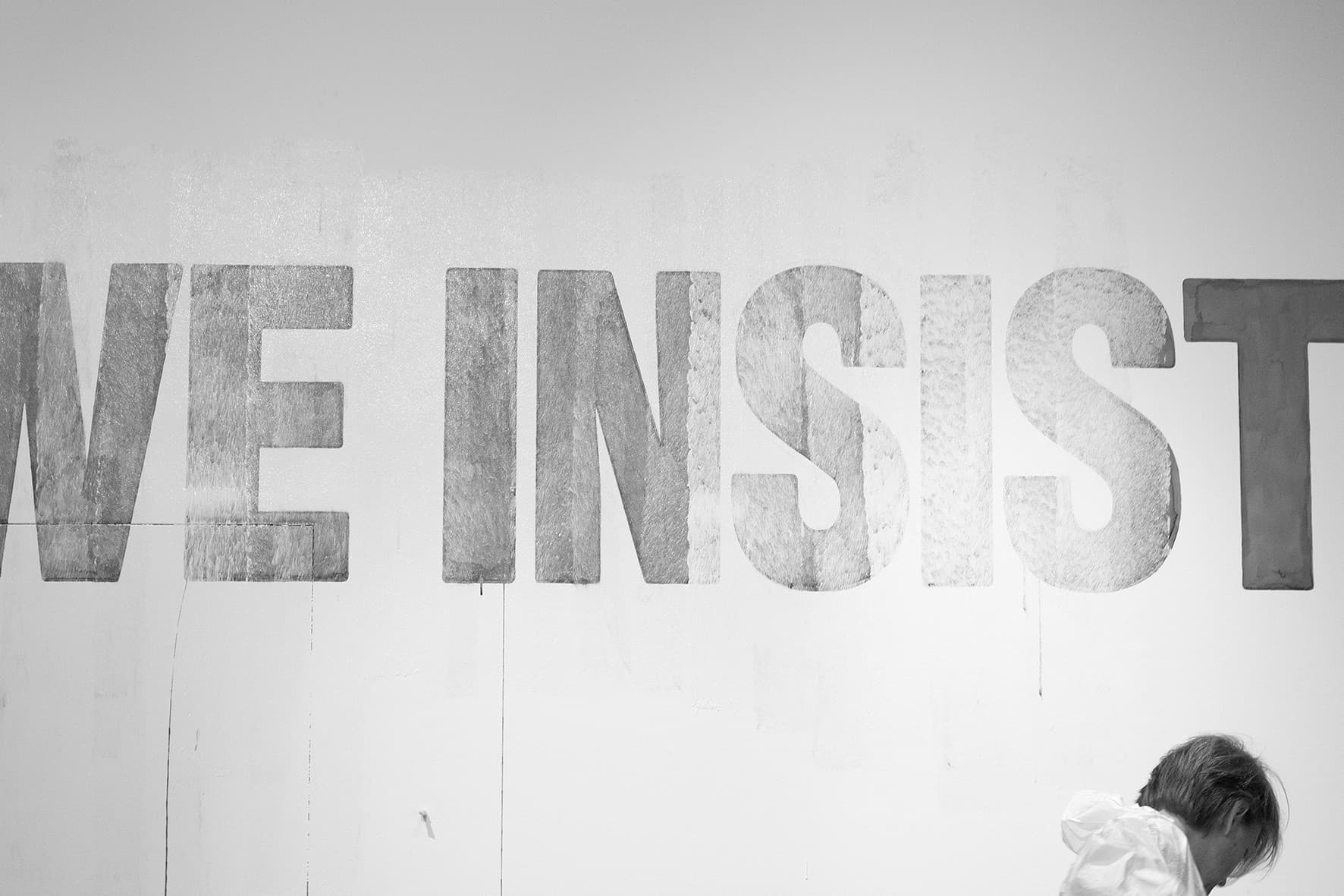Schwarz-Weiß-Aufnahme: Die Worte „WE INSIST“ in großen grauen Buchstaben auf einer weißen Wand, rechts unten ist eine Person angeschnitten.
