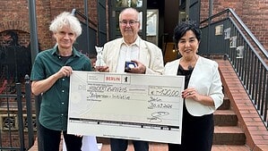 Drei Personen stehen vor dem Eingang eines Backsteingebäudes, der Mann in der Mitte hält eine Auszeichnung, vor ihnen ein großer Scheck für die Stolperstein-Initiative.