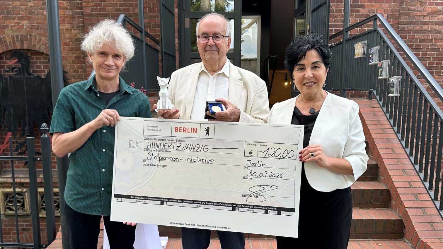 Drei Personen stehen vor dem Eingang eines Backsteingebäudes, der Mann in der Mitte hält eine Auszeichnung, vor ihnen ein großer Scheck für die Stolperstein-Initiative.