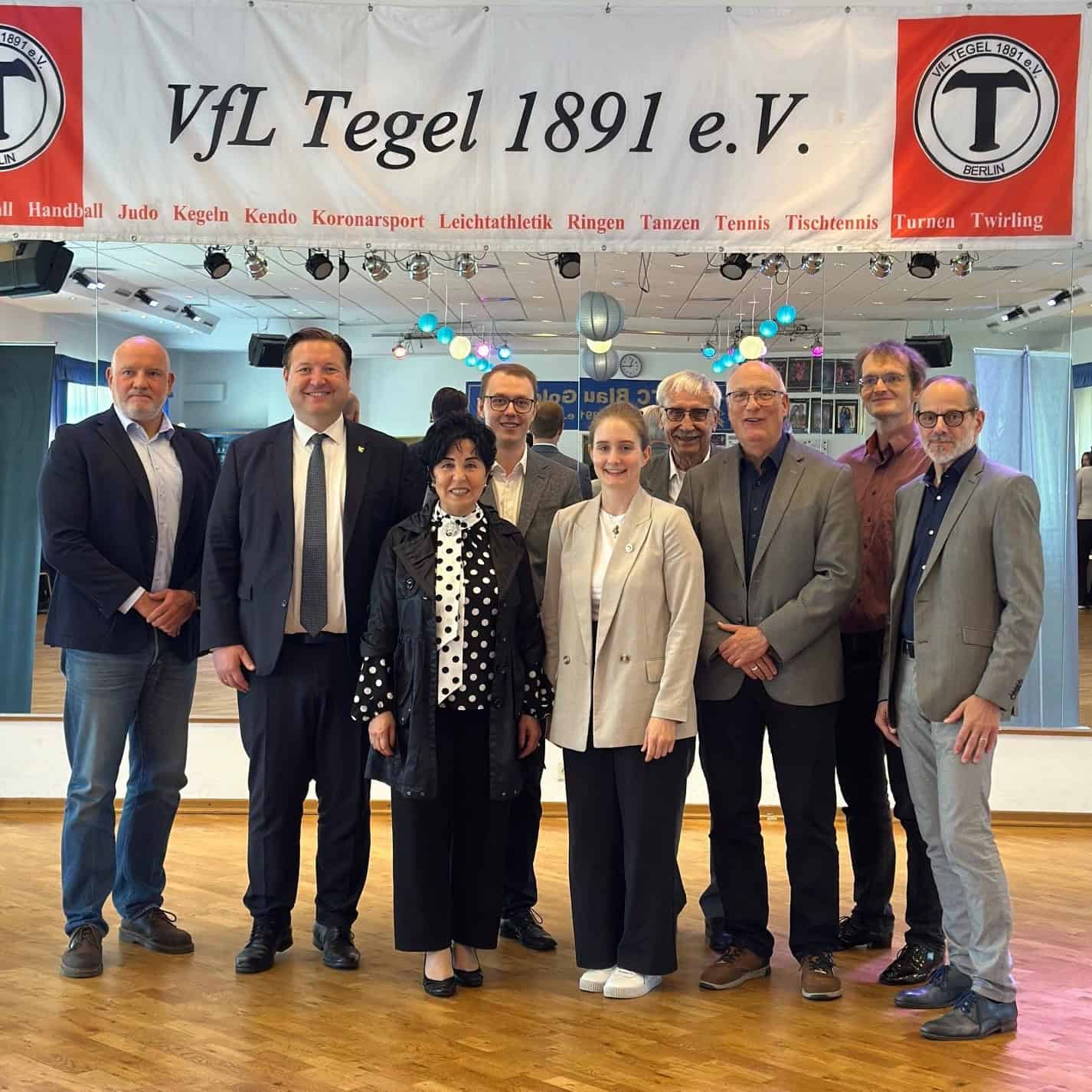 Neun Personen stehen in einem Saal mit Parkettboden nebeneinander für ein Gruppenfoto, über ihnen ein Banner mit der Aufschrift VfL Tegel 1891 e.V. und den Sportarten des Vereins.