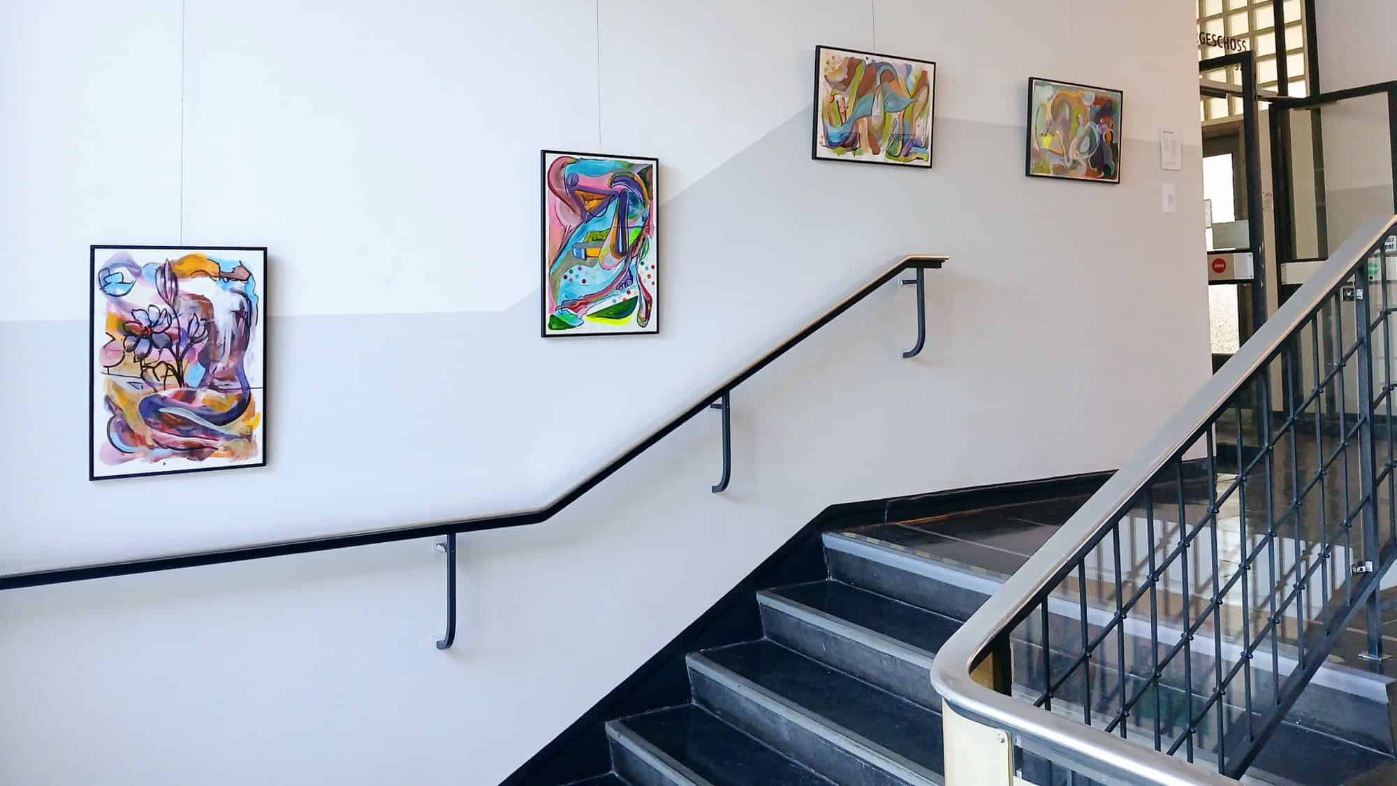 Treppenaufgang im Rathaus Reinickendorf mit gerahmten, farbenfrohen Gemälden an der weißen Wand und einem Metallgeländer.