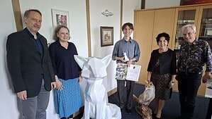 Fünf Personen stehen in einem Büro neben einer weißen Fuchsskulptur, in der Mitte ein junger Mann mit Urkunde und kleiner Fuchsfigur.