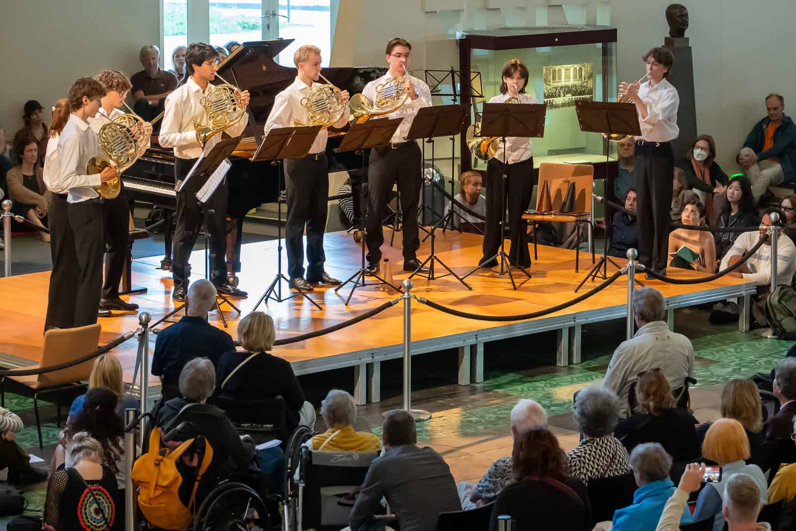 Junge Hornistinnen und Hornisten spielen auf einer Bühne im Foyer der Berliner Philharmonie vor zahlreichem Publikum.