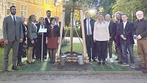 Eine größere Gruppe festlich gekleideter Menschen steht am Abend vor dem Rathaus Reinickendorf neben einem frisch gepflanzten, abgestützten Baum mit zwei Gießkannen.