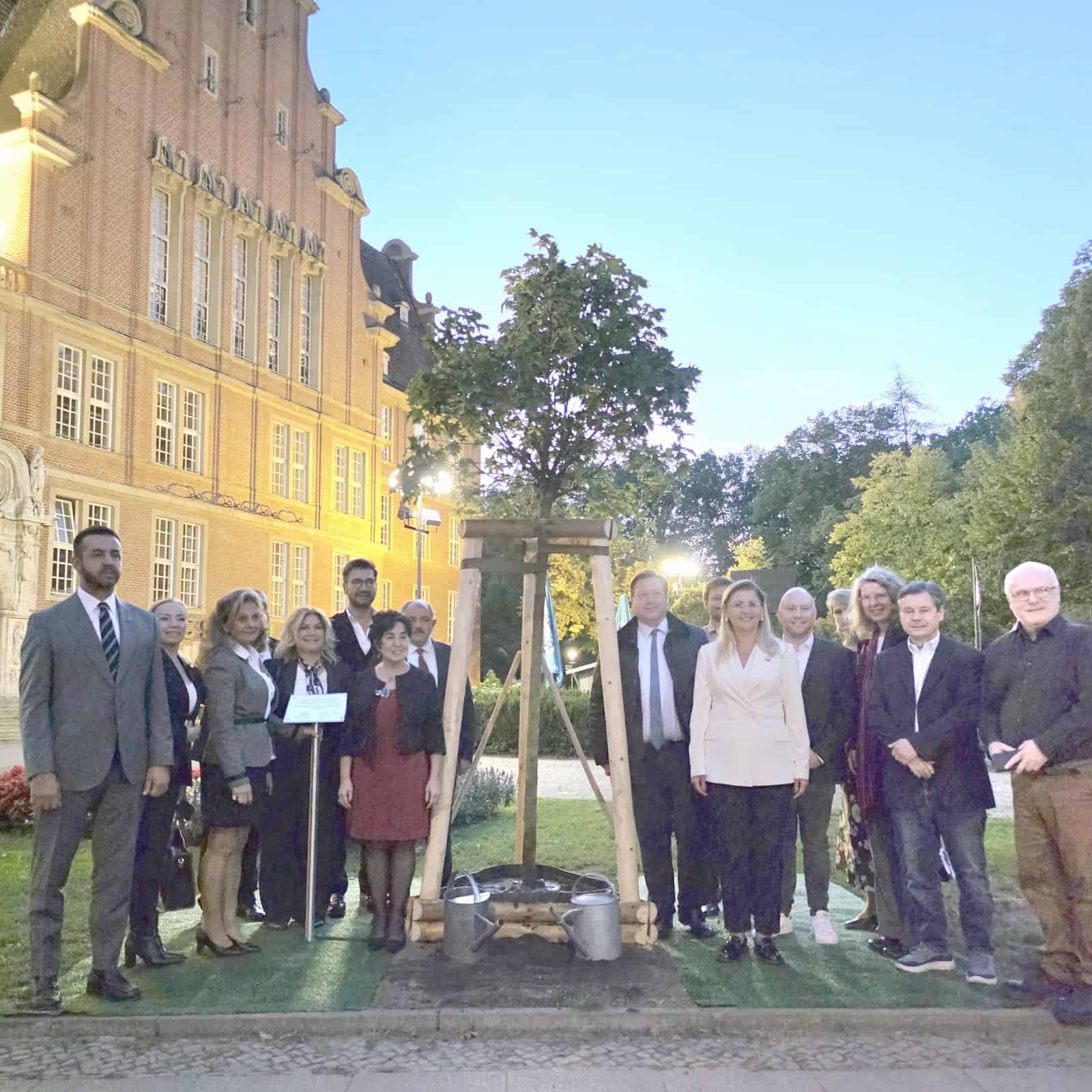 Eine größere Gruppe festlich gekleideter Menschen steht am Abend vor dem Rathaus Reinickendorf neben einem frisch gepflanzten, abgestützten Baum mit zwei Gießkannen.