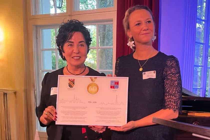 Zwei Frauen halten gemeinsam eine gerahmte Urkunde mit den Wappen von Reinickendorf und Antony und einer goldenen 60 in einem Saal mit Flügel.