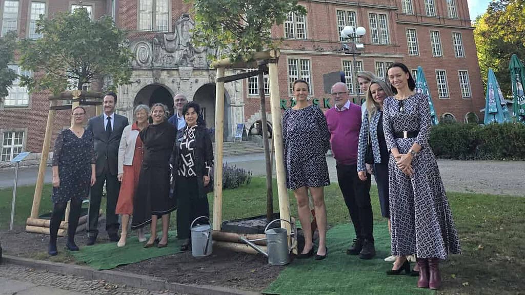 Gruppe von elf Personen neben einem frisch gepflanzten jungen Baum mit Stützpfählen und Gießkannen, im Hintergrund das Rathaus Reinickendorf mit Uhrturm.
