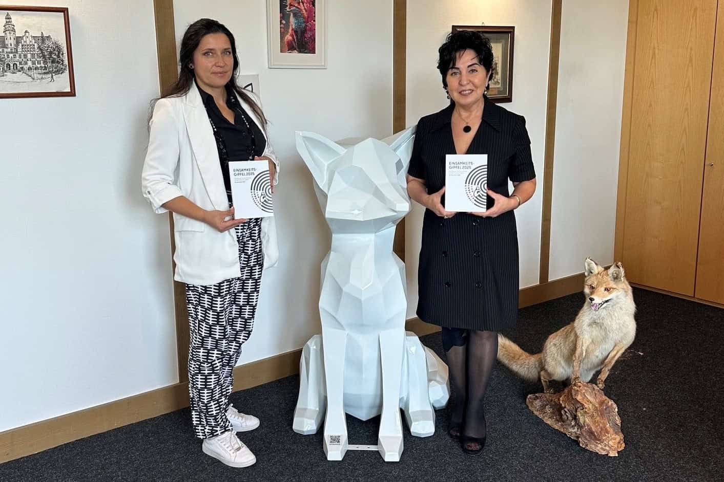 Zwei Frauen stehen in einem Büroraum neben einer weißen Fuchsskulptur und halten je ein Exemplar der Dokumentation Einsamkeitsgipfel 2025 in den Händen, auf dem Teppich davor liegen weitere Exemplare im Halbkreis, rechts ein ausgestopfter Fuchs