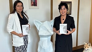 Zwei Frauen stehen in einem Büroraum neben einer weißen Fuchsskulptur und halten je ein Exemplar der Dokumentation Einsamkeitsgipfel 2025 in den Händen, auf dem Teppich davor liegen weitere Exemplare im Halbkreis, rechts ein ausgestopfter Fuchs