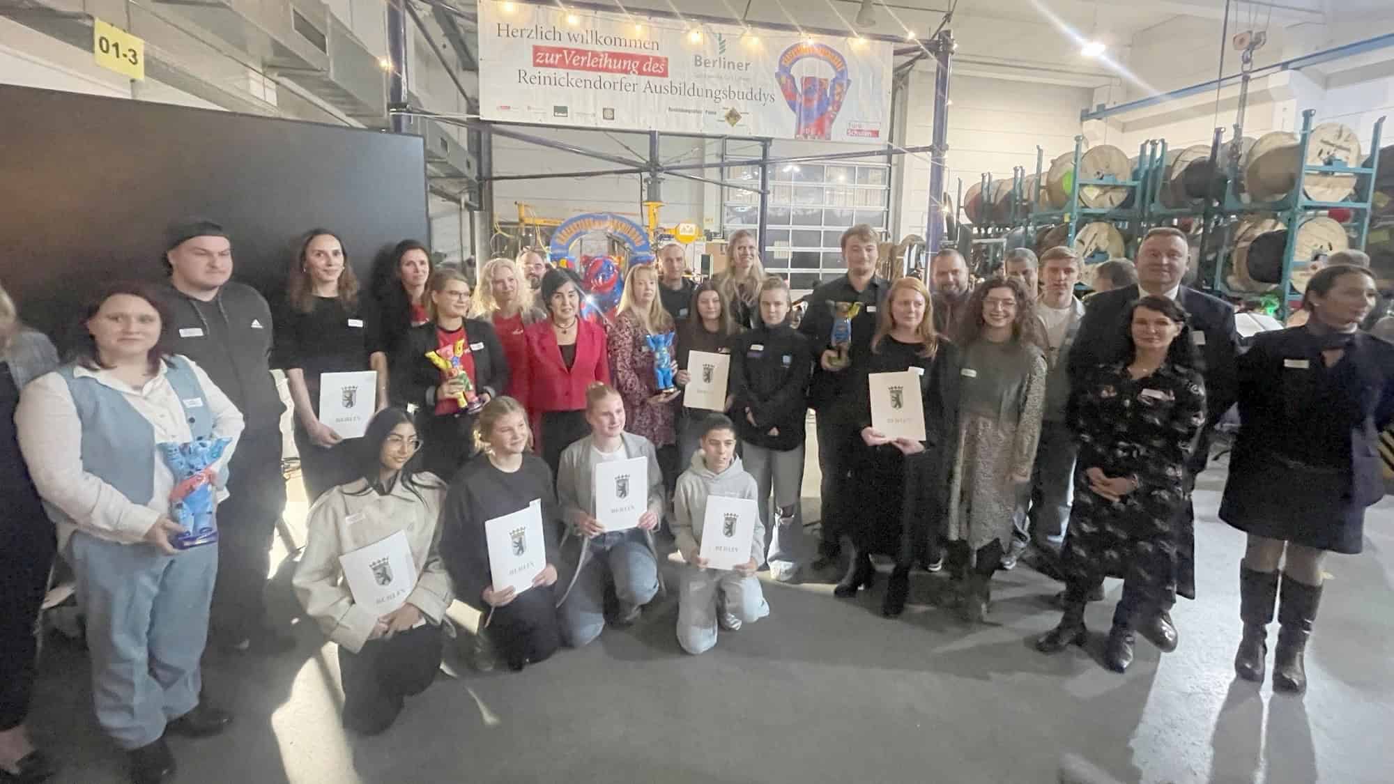 Gruppenfoto in einer Werkshalle mit Kabeltrommeln: Rund 30 Menschen, einige mit Urkunden und bunten Bären-Trophäen, stehen und knien vor einem Banner zur Verleihung des Reinickendorfer Ausbildungsbuddys