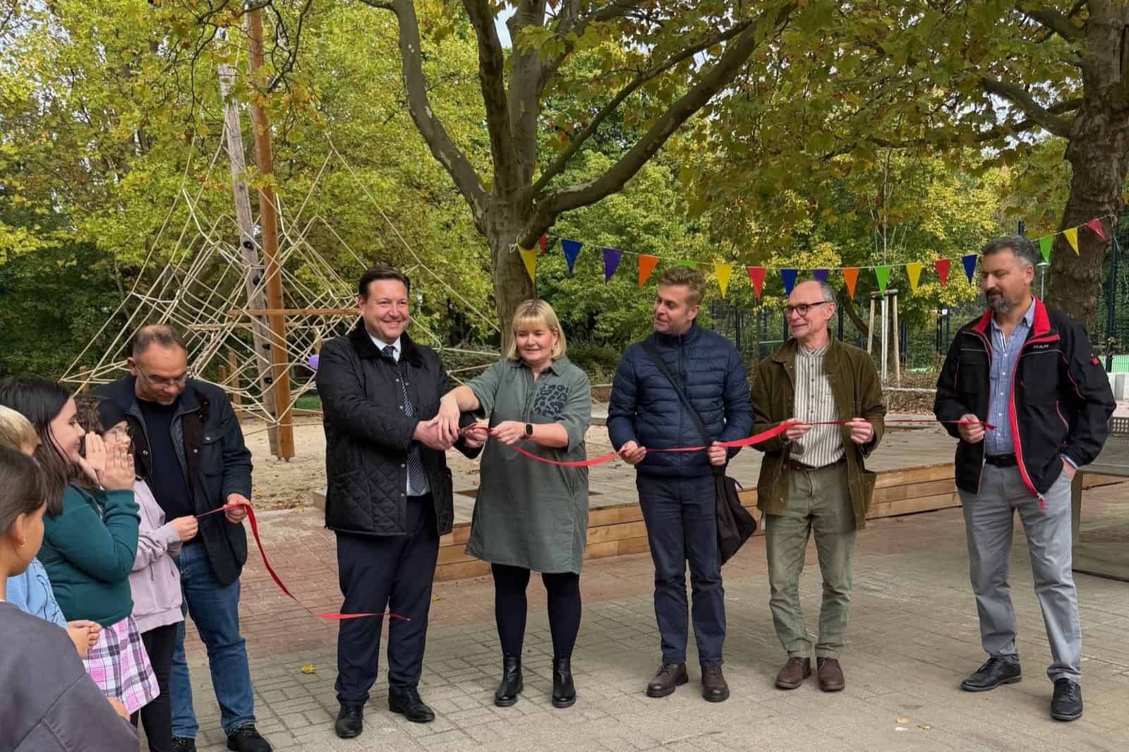 Sechs Erwachsene stehen mit einem roten Band auf einem gepflasterten Schulhof, dahinter ein Klettergerüst und bunte Wimpel, links schauen Kinder zu.