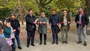 Sechs Erwachsene stehen mit einem roten Band auf einem gepflasterten Schulhof, dahinter ein Klettergerüst und bunte Wimpel, links schauen Kinder zu.
