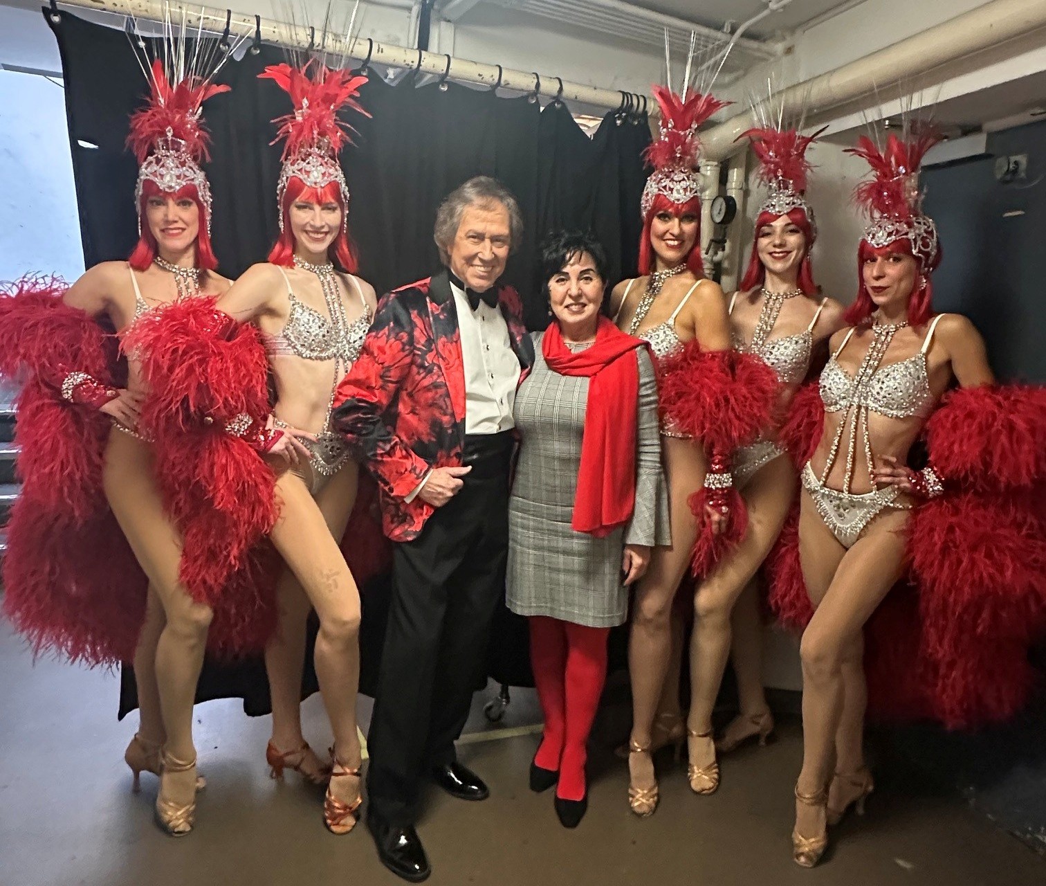 Bezirksbürgermeisterin Emine Demirbüken-Wegner und Entertainer Bert Beel im rot-schwarzen Sakko zwischen fünf Can-Can-Tänzerinnen mit roten Federkostümen und Kopfschmuck.